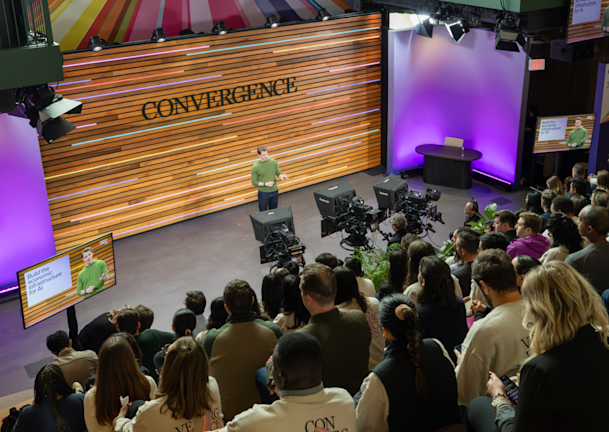 Speaker on stage at Stripe's Convergence event addressing a seated audience, with 'Build the economic infrastructure for AI' displayed on monitors.