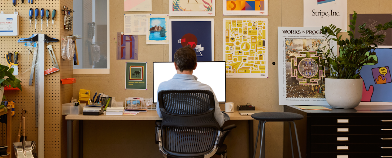 Person at a desk in a Stripe office workspace with design prints, hand tools on a pegboard, and two monitors.