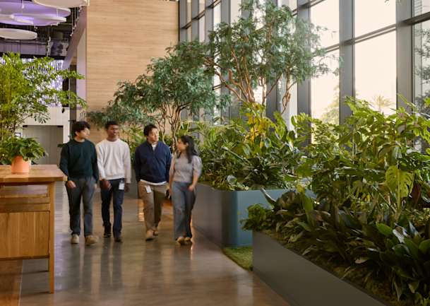 Four coworkers walk through a sunlit office atrium filled with large indoor plants.